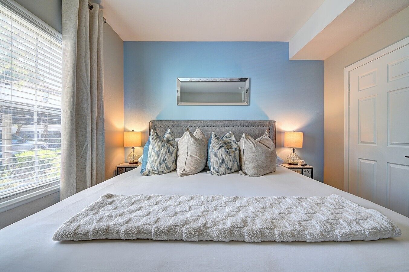 A bedroom with a bed, a headboard, bedside lamps, a window with blinds, and a door.