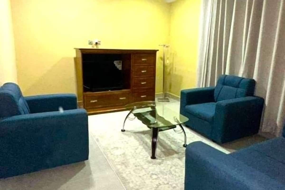 A living room with a flat-screen TV, two blue sofas, a glass coffee table, a ceiling fan, and a large window with curtains.