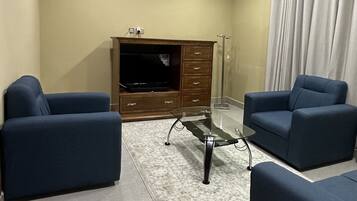 Un soggiorno con due divani blu, un tavolino in vetro, un mobiletto in legno con una TV a schermo piatto e una ventola a soffitto.
