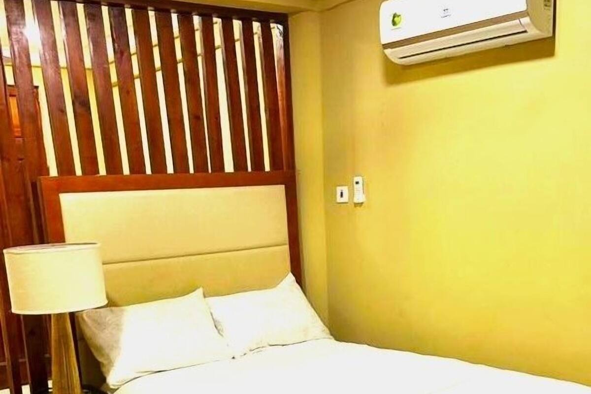 A hotel room with a bed, nightstand, lamp, wooden headboard, air conditioning unit, and tiled floor.