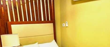 A hotel room with a bed, nightstand, lamp, wooden headboard, air conditioning unit, and tiled floor.
