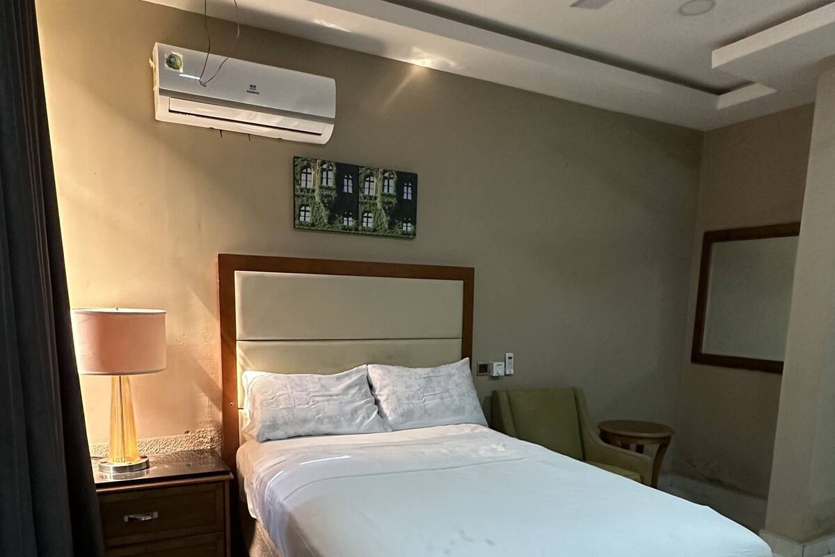 A modern hotel room with a large bed, a ceiling fan, an air conditioning unit, a bedside lamp, and a small table.