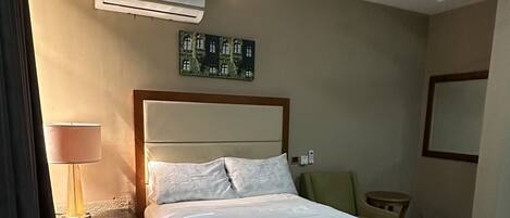 A modern hotel room with a large bed, a ceiling fan, an air conditioning unit, a bedside lamp, and a small table.