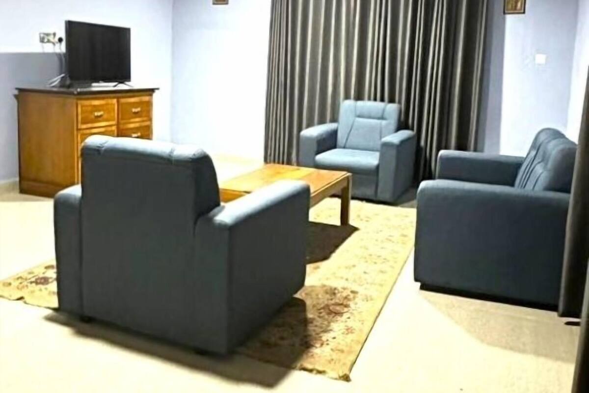 A living room with a flat-screen TV, a wooden cabinet, a coffee table, two armchairs, and a large window with curtains.