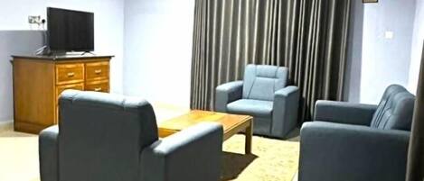 A living room with a flat-screen TV, a wooden cabinet, a coffee table, two armchairs, and a large window with curtains.