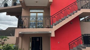 A two-story building with a red facade, a balcony with decorative railings, and a staircase with ornate balusters. - ACHIE SUITES (Accra)