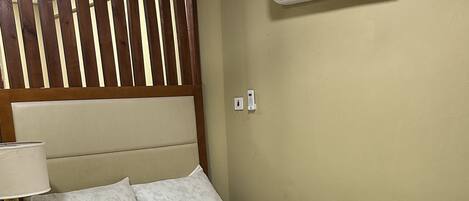 A neatly made bed with a headboard, a bedside table with a lamp, and an air conditioning unit on the wall.