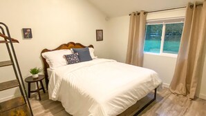 A neatly made bed with white linens and a wooden headboard, a small bedside table with a plant, and a window with beige curtains. - The Modern Cottage in Rural Lake Stevens (Lake Stevens)