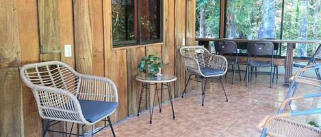 A covered outdoor seating area with wicker chairs and a small table, wooden walls, and large windows.