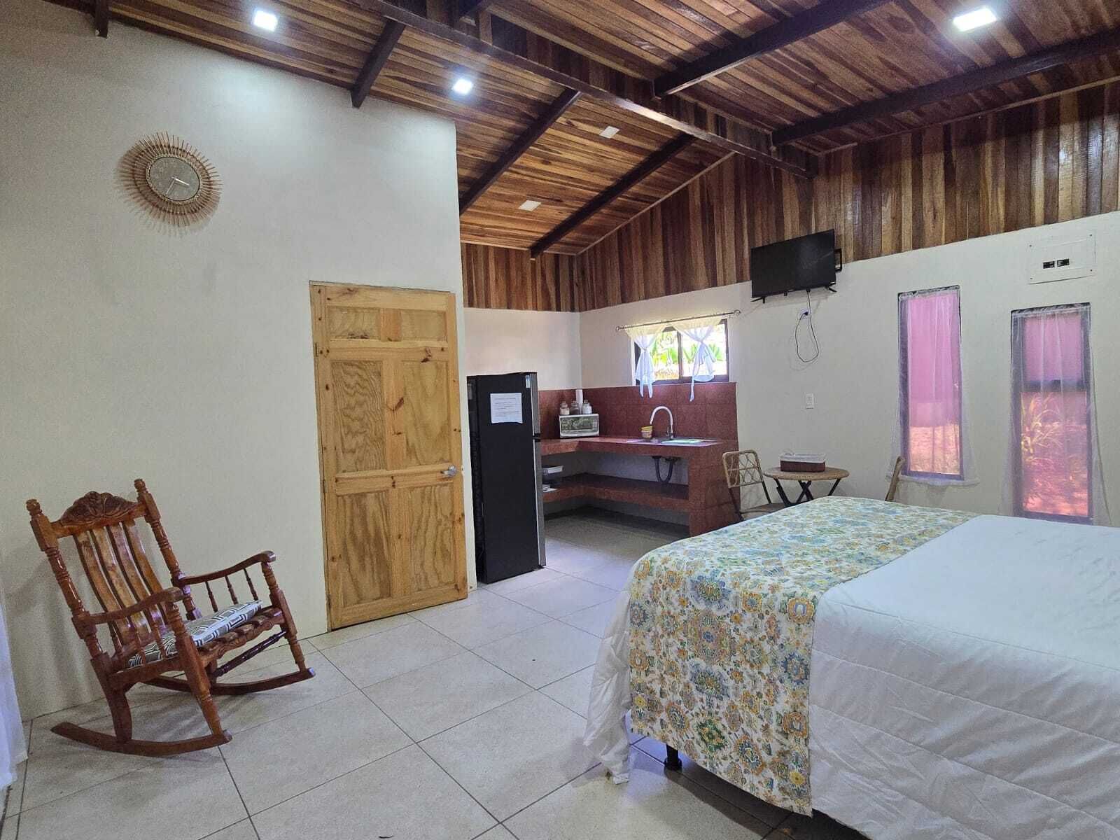 A room with a bed, a wooden rocking chair, a desk with a sink, and a television on the wall.