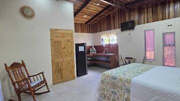 A room with a bed, a wooden rocking chair, a desk with a sink, and a television on the wall.