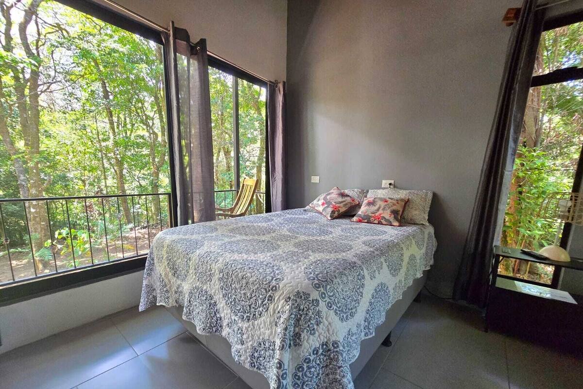 A bedroom with a large bed, a view of trees through the window, and a balcony.