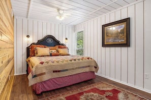 A bedroom with a wooden headboard, a quilted bedspread, a checkered skirt, a patterned rug, and a framed landscape painting on the wall.