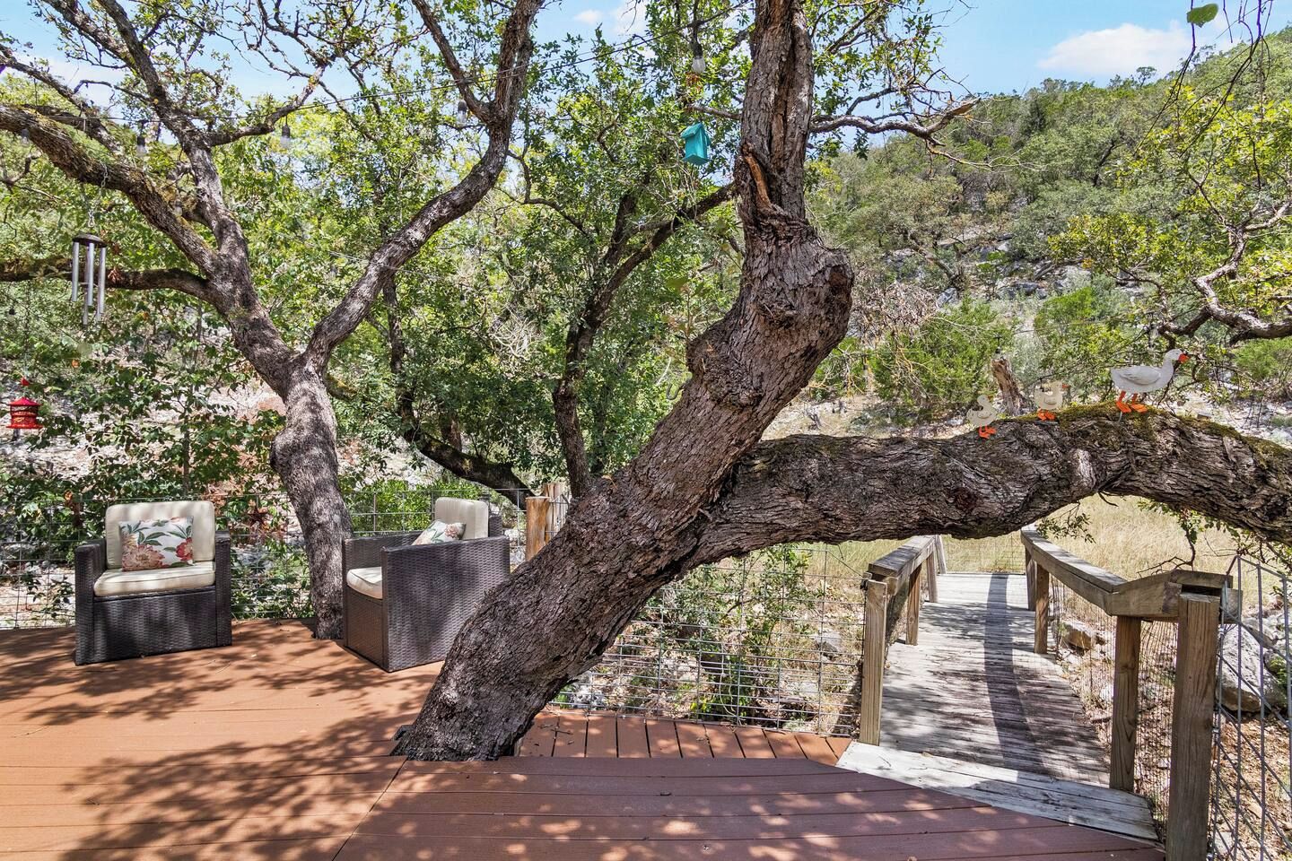 Eine Holzterrasse mit Korbmöbeln, ein Baum mit schräg stehendem Stamm und eine Holzbrücke.