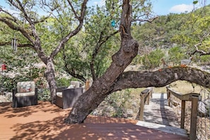 Eine Holzterrasse mit Korbmöbeln, ein Baum mit schräg stehendem Stamm und eine Holzbrücke.