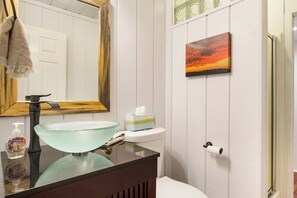 Une salle de bain avec un lavabo en verre, un meuble-lavabo en bois, un miroir, des toilettes et un tableau accroché au mur.