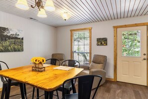 A dining area with a wooden table, chairs, and a vase of sunflowers. There are windows with blinds, a door, and wall-mounted artwork. - Big Fern | Pet-Friendly Cabin Near Frio & Garner (Leakey)