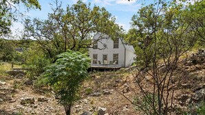 A white house with a porch, surrounded by trees and rocky terrain. - Big Fern Cabin in Leakey (Leakey)