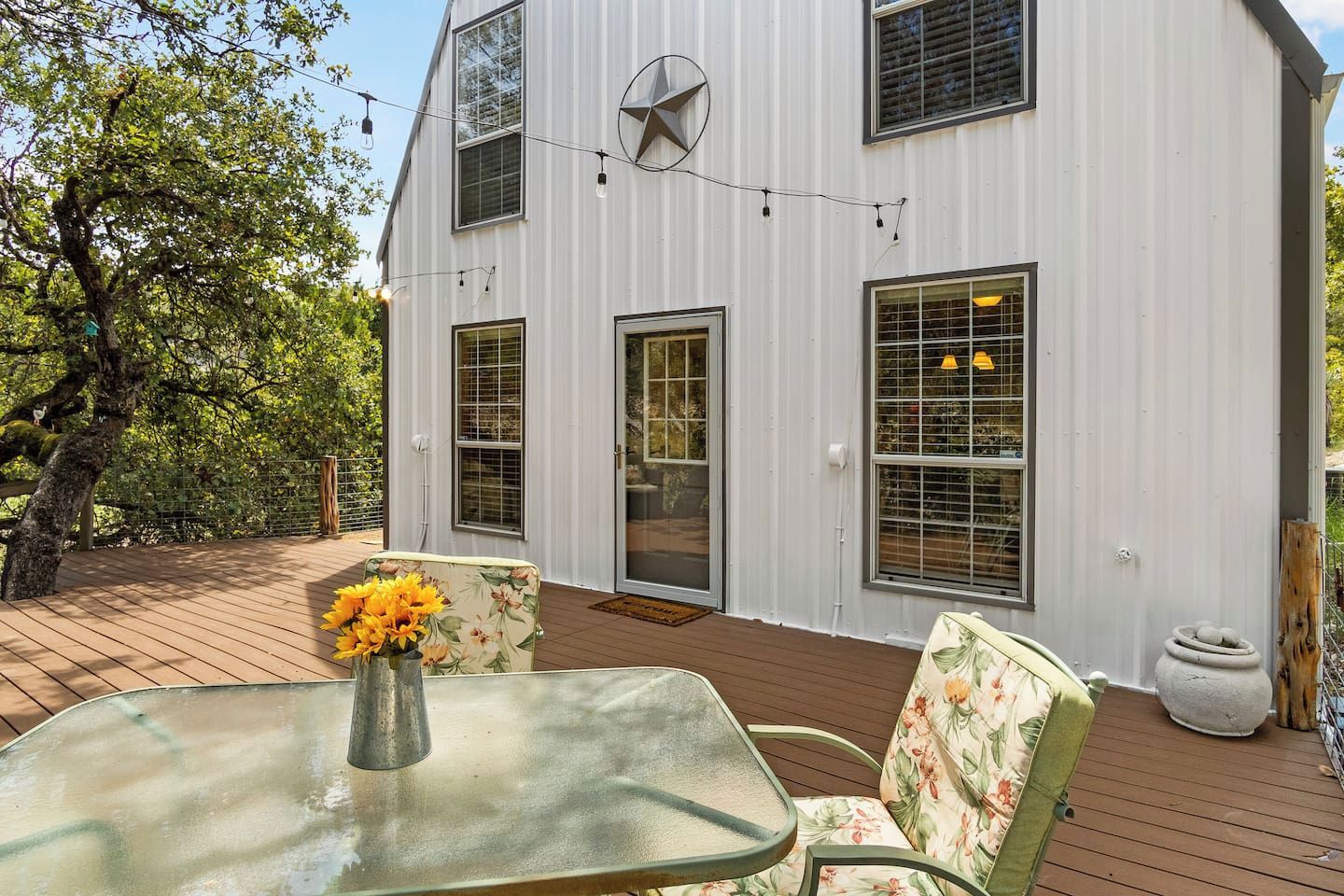 A white building with a large window and a door, a deck with chairs and a table, and a tree in the background.