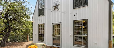 A white building with a large window and a door, a deck with chairs and a table, and a tree in the background.