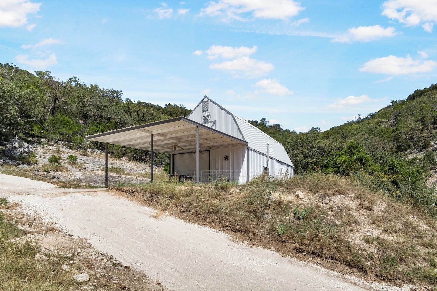 Edifício térreo com varanda coberta e porta de garagem, cercado por paisagem natural e estrada de terra.