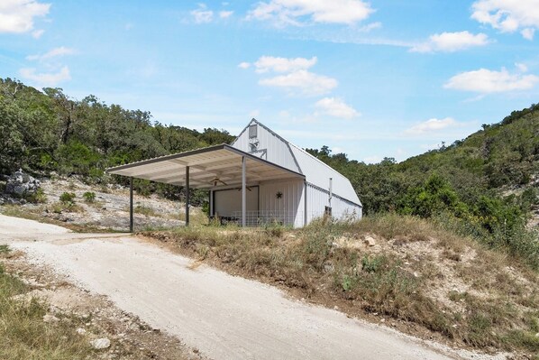 A single-story building with a covered porch and a garage door, surrounded by natural landscape and a dirt road.