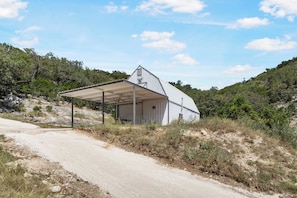 A single-story building with a covered porch and a garage door, surrounded by natural landscape and a dirt road. - Big Fern | Pet-Friendly Cabin Near Frio & Garner (Leakey)