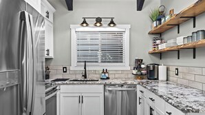 A modern kitchen with white cabinets, stainless steel appliances, a window with blinds, and a marble countertop. - Townhouse in Private Lakeside Community (Sandpoint)