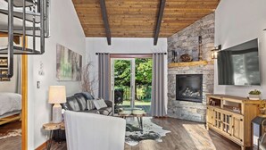 A spacious living room with a stone fireplace, a white sofa, and a TV mounted on the wall. - Townhouse in Private Lakeside Community (Sandpoint)