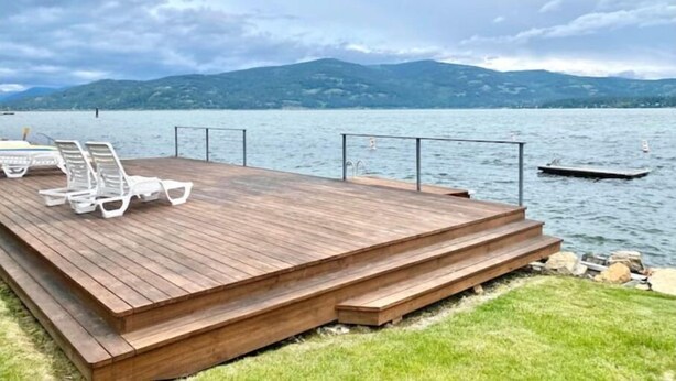 A wooden deck with white lounge chairs and a pool, overlooking a lake with mountains in the background. - Townhouse in Private Lakeside Community (Sandpoint)