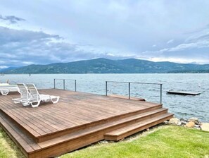 A wooden deck with white lounge chairs and a pool, overlooking a lake with mountains in the background. - Townhouse in Private Lakeside Community (Sandpoint)