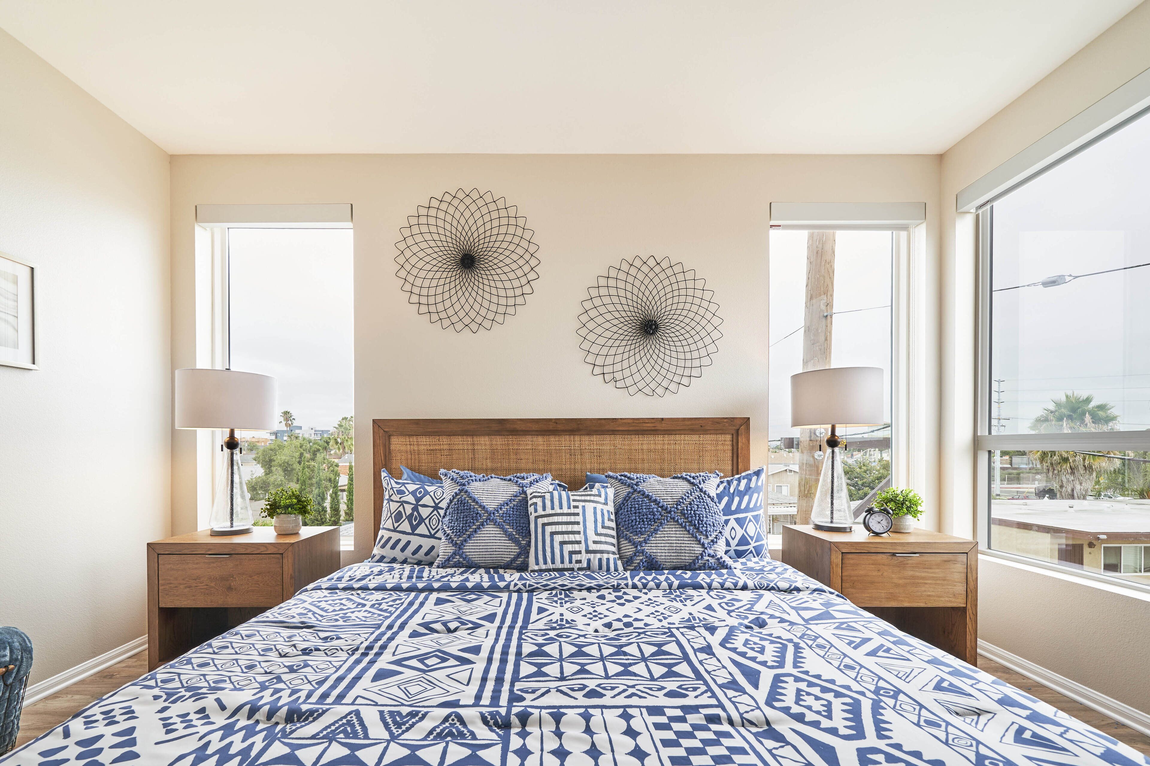A bedroom with a large bed, two wall art pieces, a wooden headboard, two bedside tables with lamps, and a view of the city through large windows.