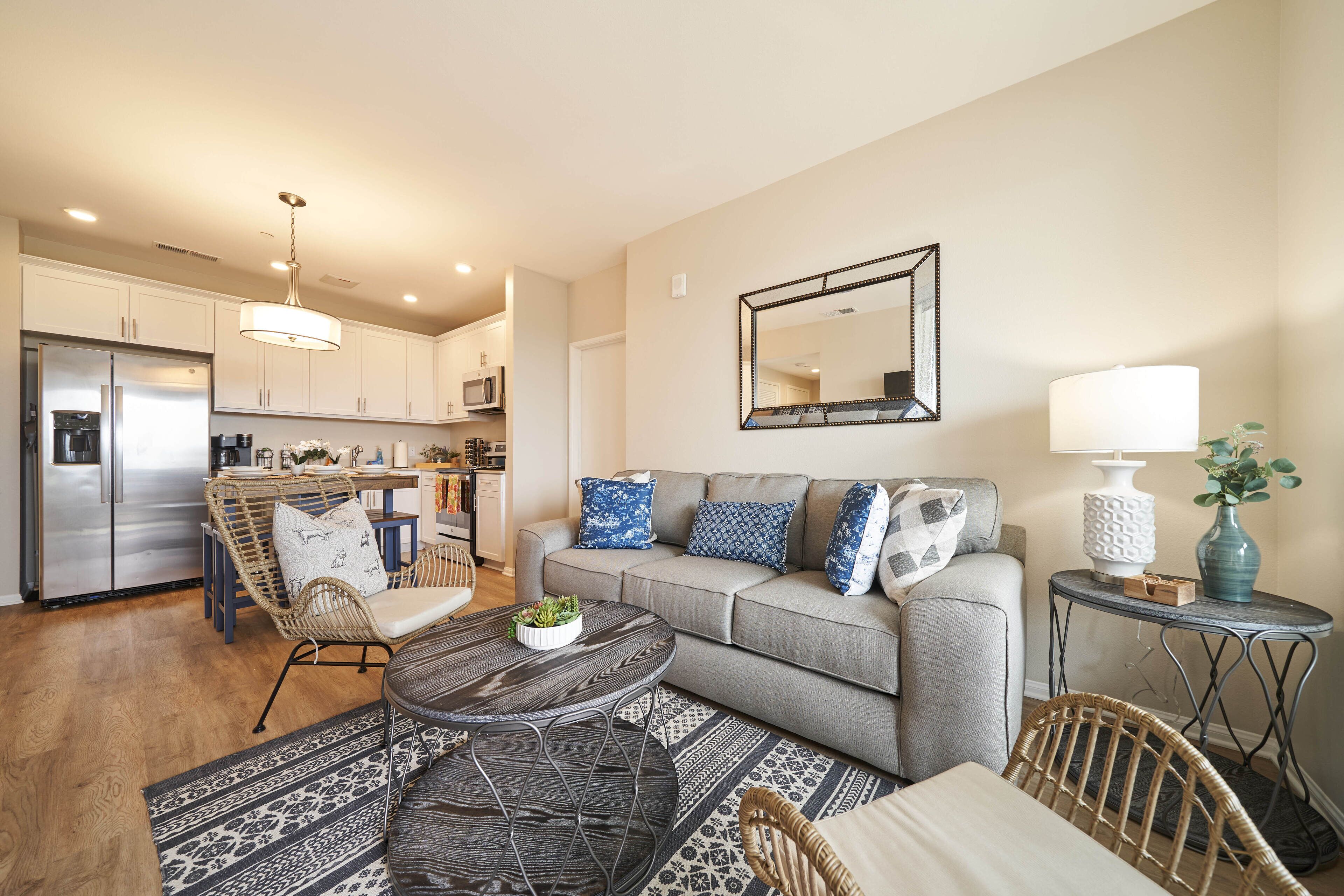 A modern living room with a grey sofa, a coffee table, wicker chairs, and a kitchen in the background.