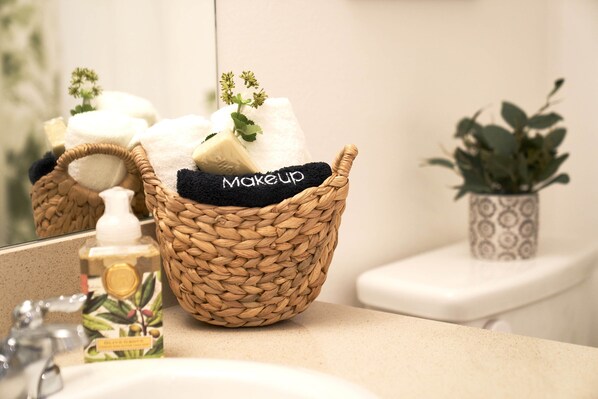 A bathroom with a woven basket containing a towel and a bar of soap, a small potted plant, and a bottle of liquid soap on the counter. - Luxury 2BD 2BA, BRAND NEW/PETS/CLOSE TO EVERYTHING/BOHO+COZY VIBES (San Diego)