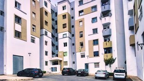 A multi-story residential building with a central entrance, several balconies, and a parking area with cars.