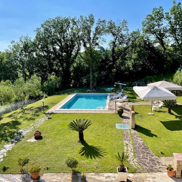 A rectangular swimming pool surrounded by a well-maintained lawn, with a stone pathway leading to a seating area with umbrellas.