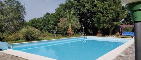 Piscina rectangular amb una escala a un costat, envoltada de vegetació i arbres frondosos.
