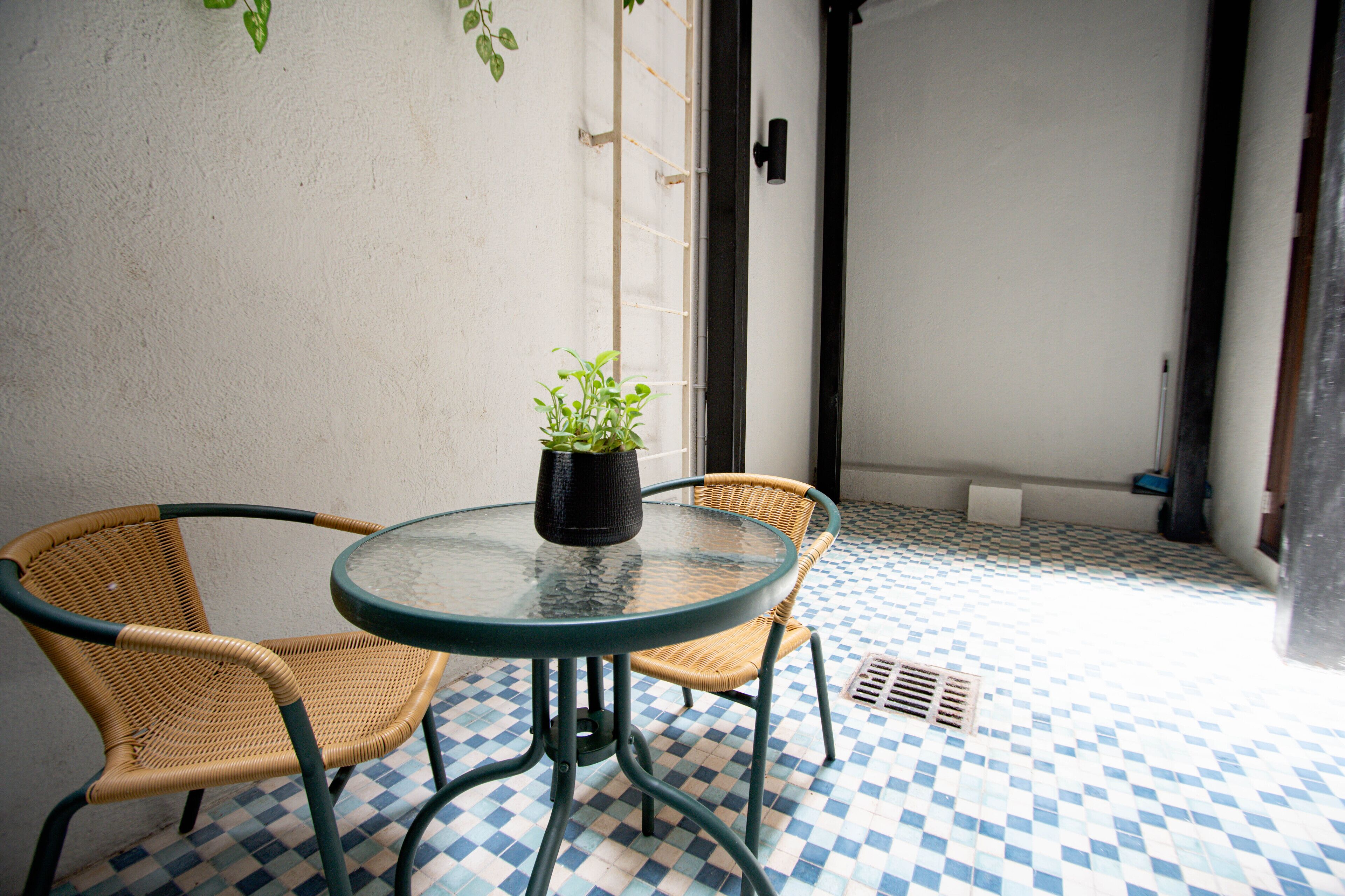 Une petite table ronde, deux chaises en osier, une plante en pot et un sol à carreaux.