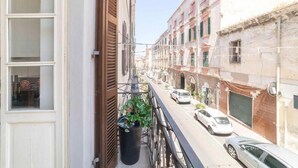 Eine enge Straße mit Häusern an beiden Seiten, ein Balkon mit einem Blumentopf und geparkte Autos.