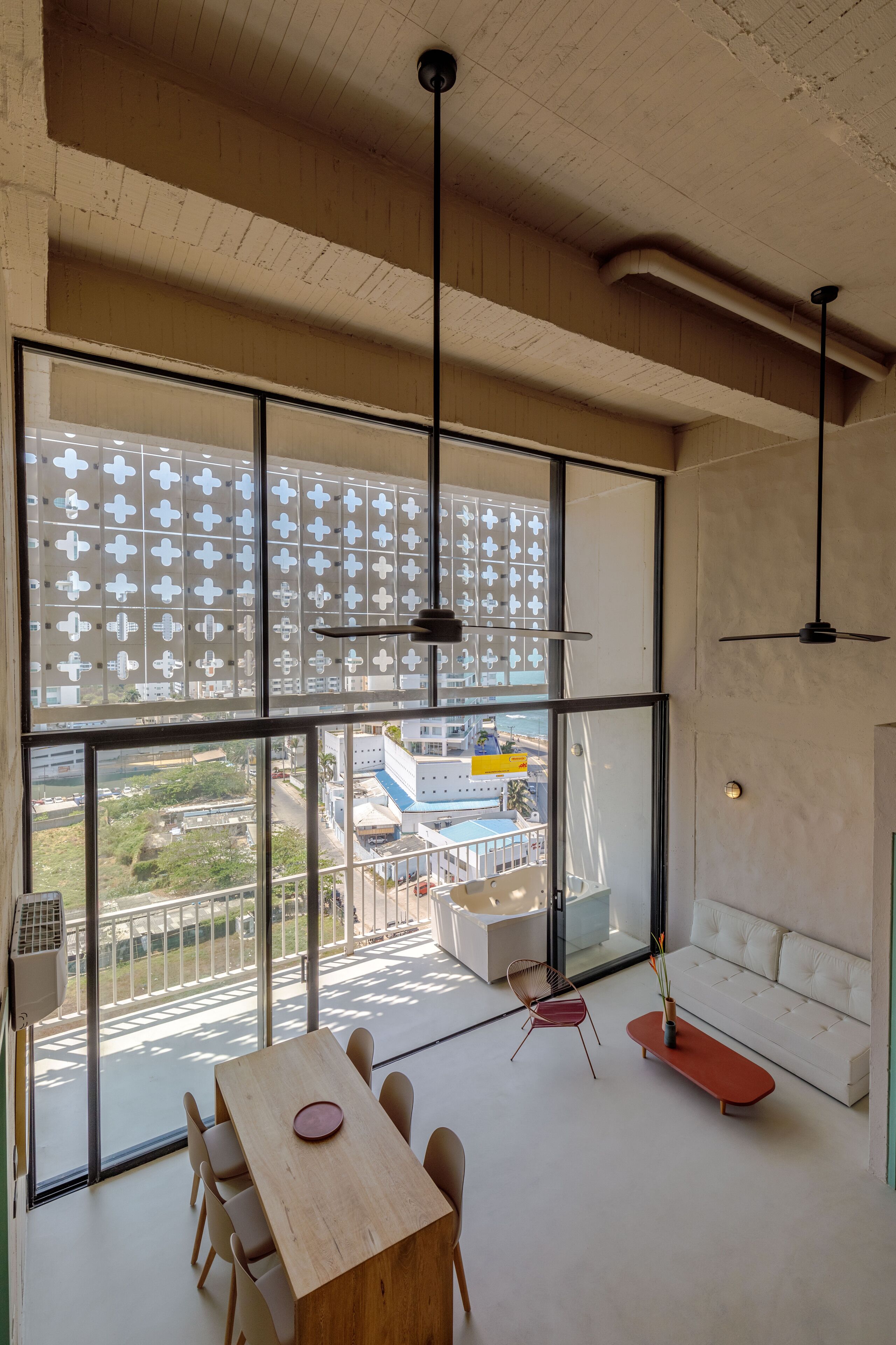 Una habitación amplia con ventanas grandes, una mesa de madera, una silla roja, un sofá blanco y un banco rojo.
