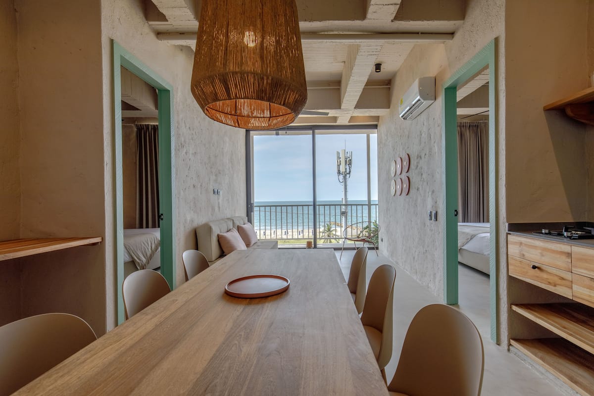 A modern dining area with a long wooden table, beige chairs, a woven pendant light, and a view of the sea through large windows.