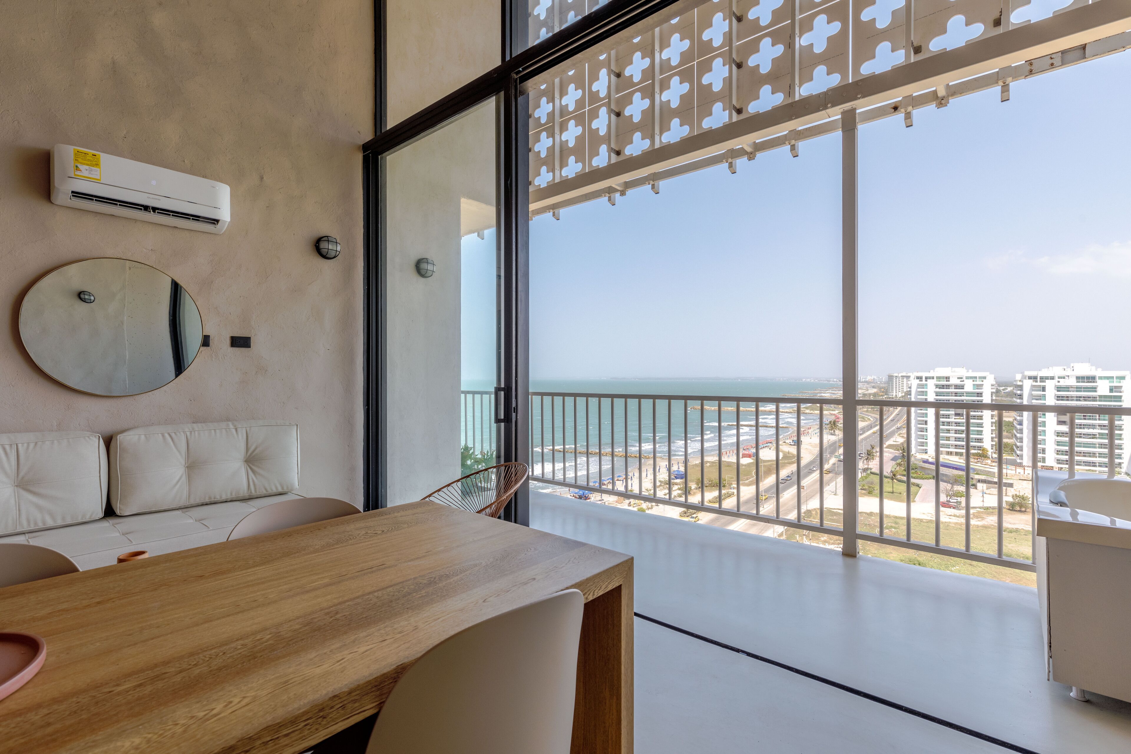 A modern balcony with a view of the beach and cityscape, a round mirror, and a wooden table.