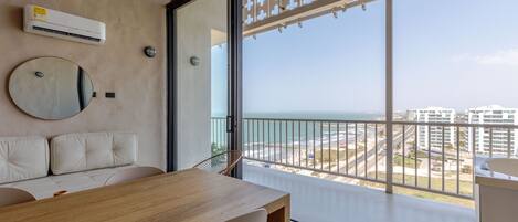 A modern balcony with a view of the beach and cityscape, a round mirror, and a wooden table.