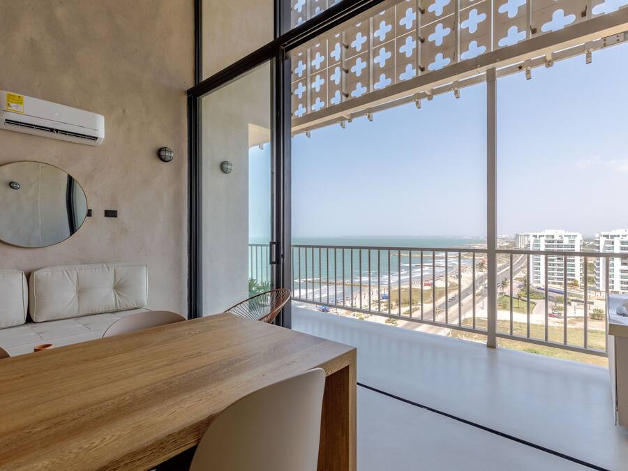 A modern balcony with a view of the beach and cityscape, a round mirror, and a wooden table.
