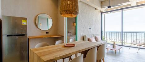 A modern kitchen with a wooden table, chairs, a refrigerator, a hanging light fixture, and a view of the sea through large windows.