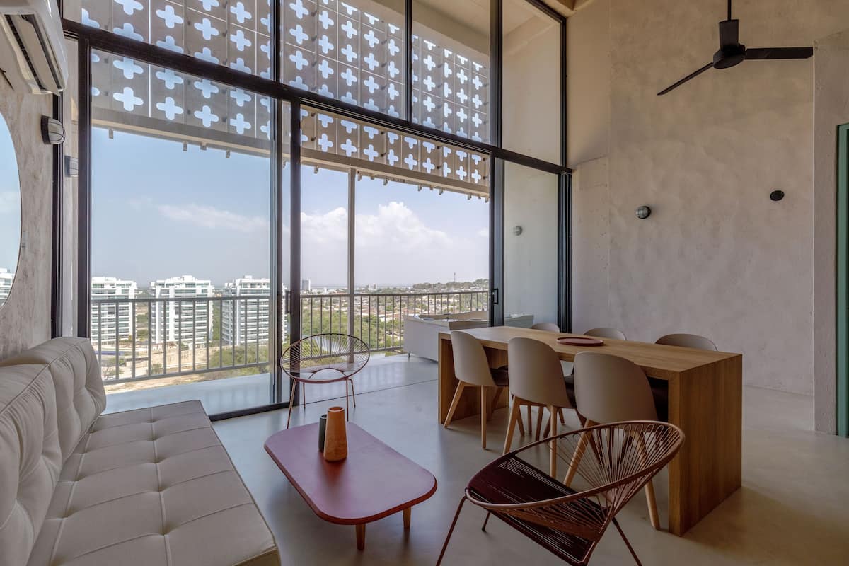 A modern living room with a large window, a sofa, a wooden table, and a dining area with chairs.