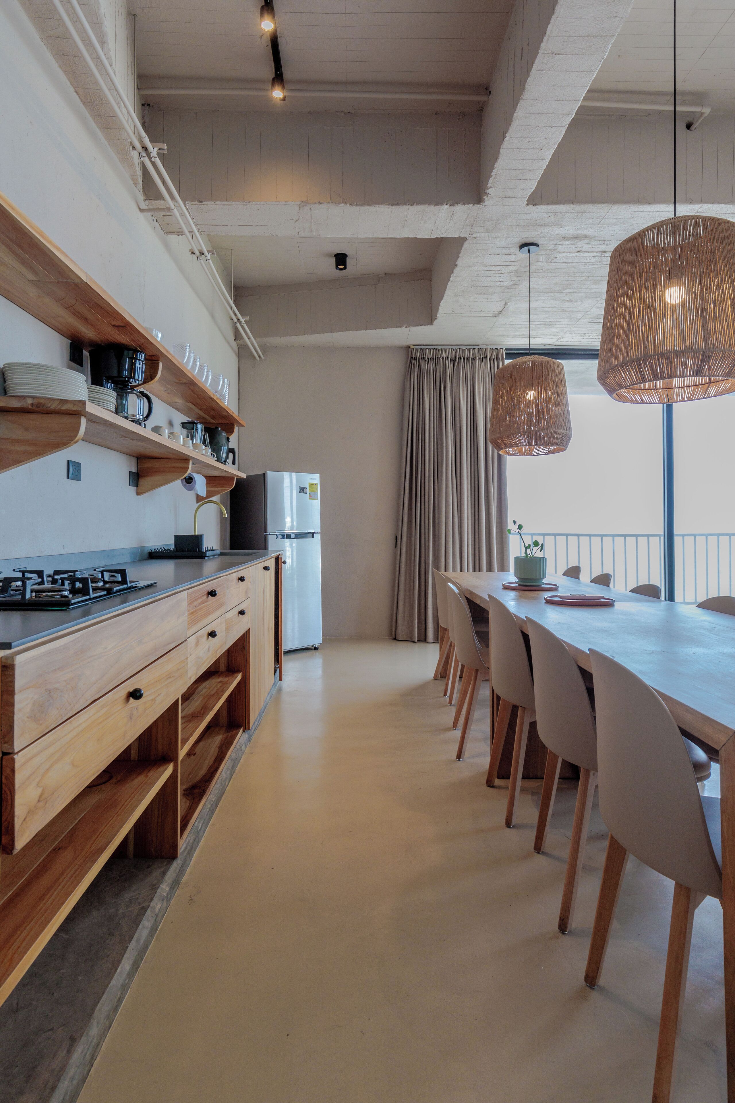 A modern kitchen with wooden cabinets, a stainless steel refrigerator, and a dining area with a long table and chairs.