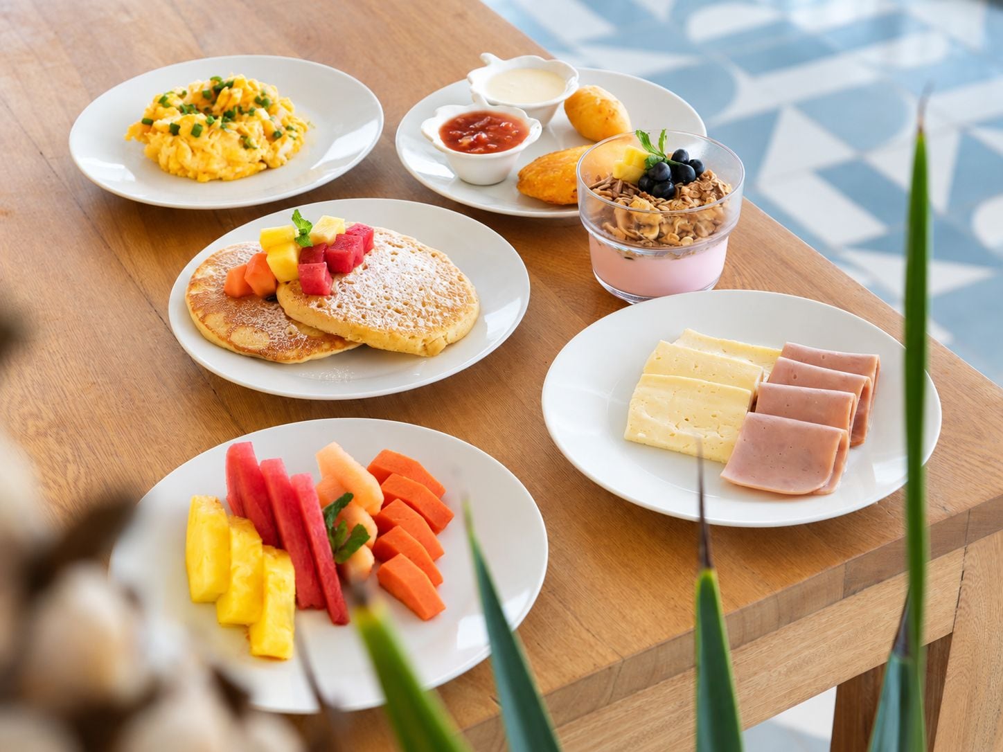 Disposição de café da manhã com ovos mexidos, panquecas com frutas, iogurte com granola, presunto e frutas frescas.