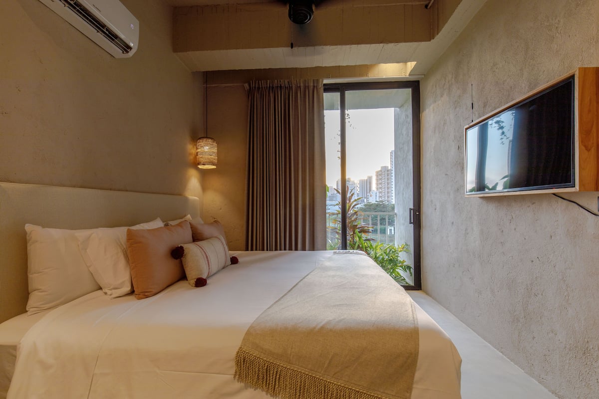 A hotel room with a large bed, a flat-screen TV mounted on the wall, and a view of the city through the window.