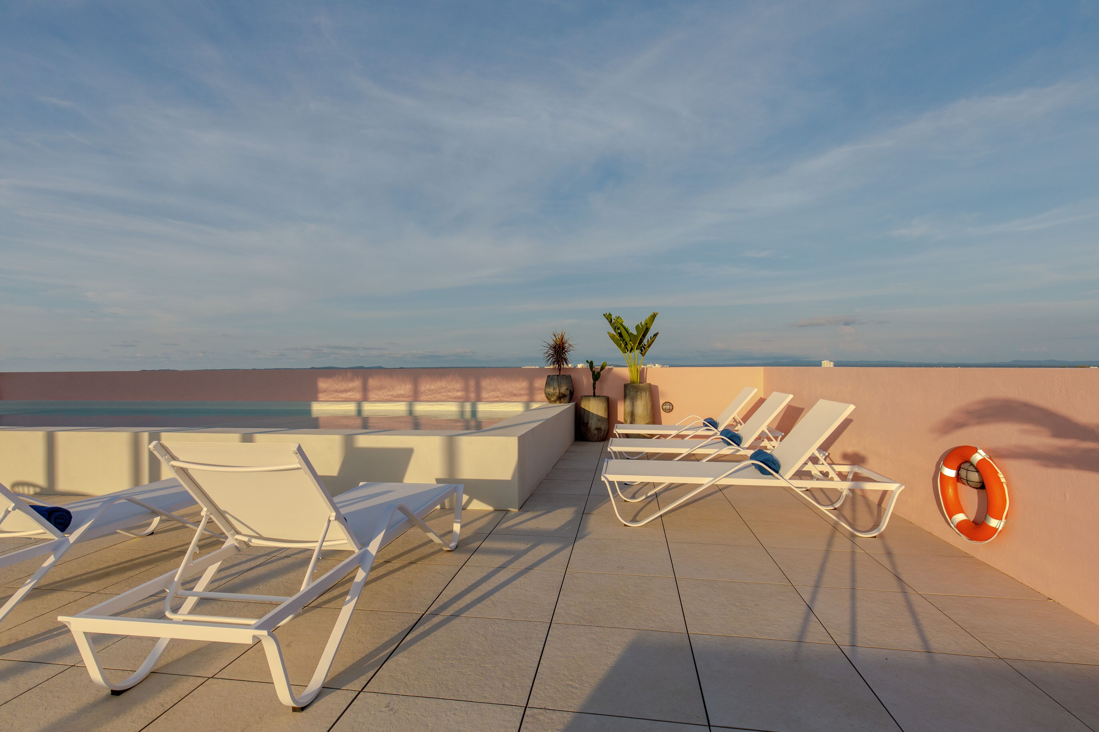 A rooftop lounge area with white sun loungers, a life ring, and potted plants.